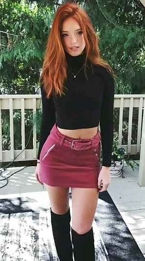 a red head woman in a black top and a pink skirt and boots standing on a porch with trees in the background